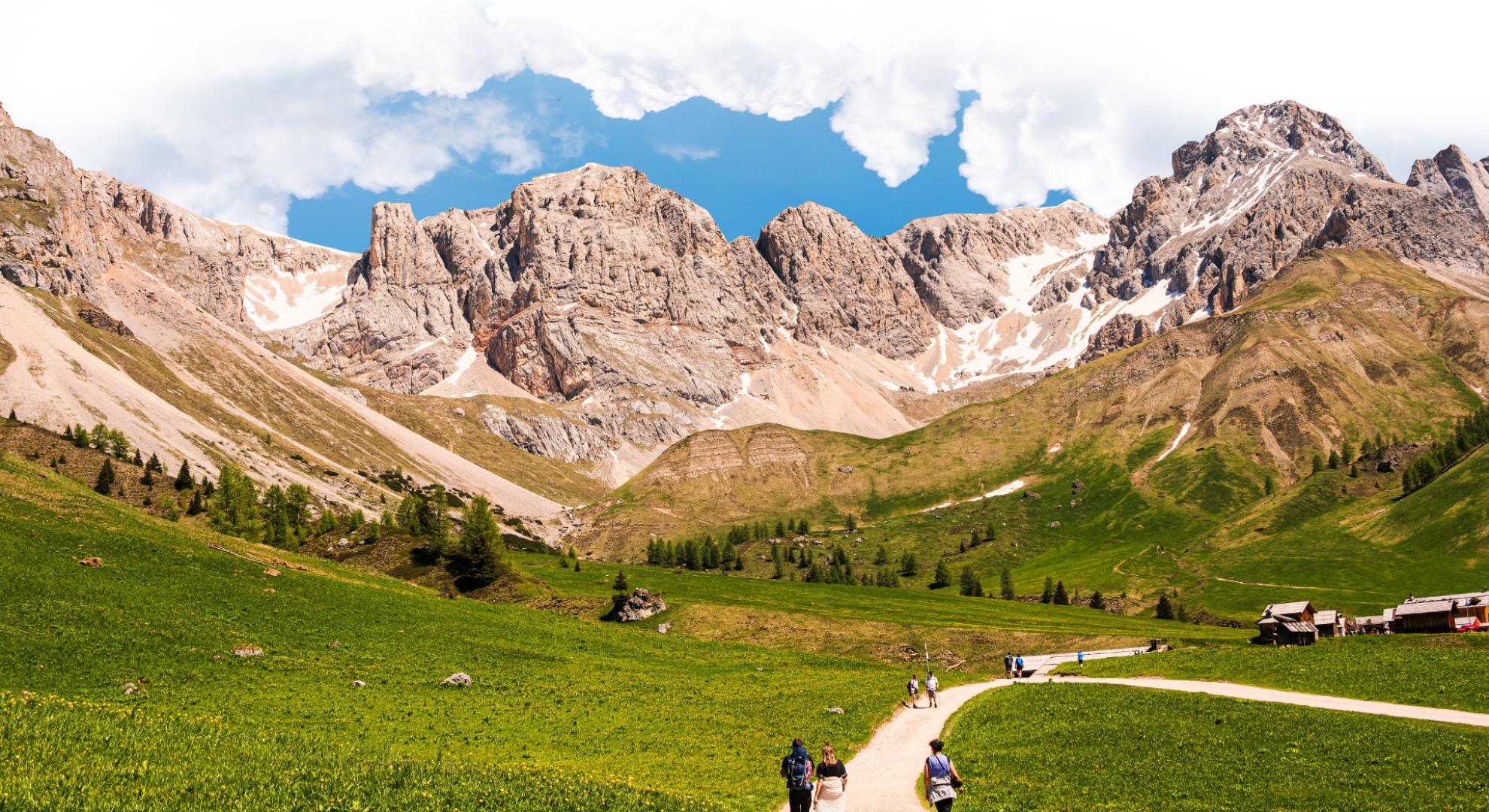 Escursionisti camminano su un sentiero di montagna tra prati verdi e cime rocciose.